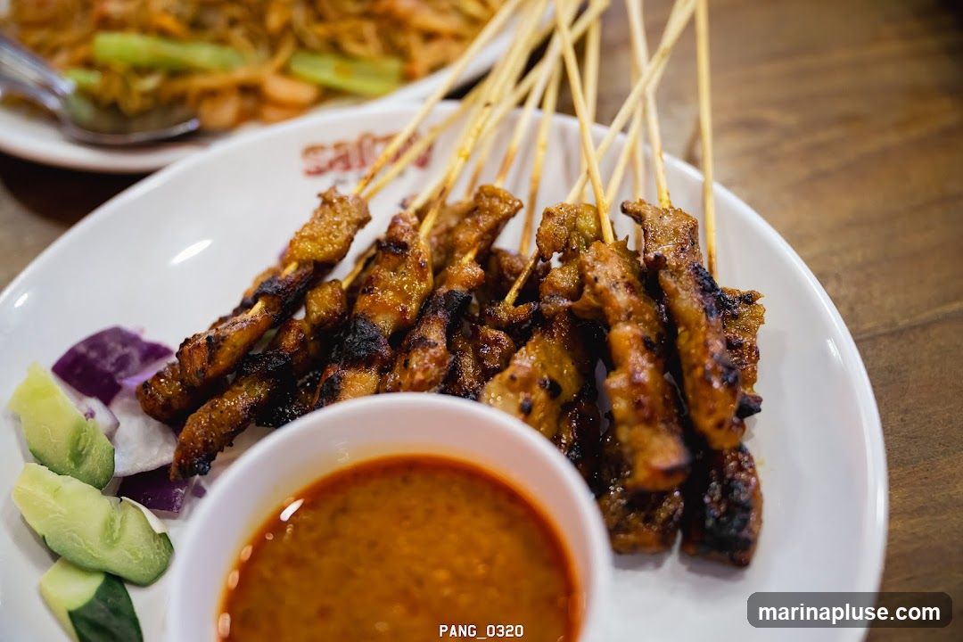 A close-up shot of grilled satay skewers served on a white plate with cucumber slices, red onion, and a small bowl of peanut sauce. In the background, another plate of food, possibly noodles, is visible. The restaurant's name, "Sajian," is partially visible on the plate.