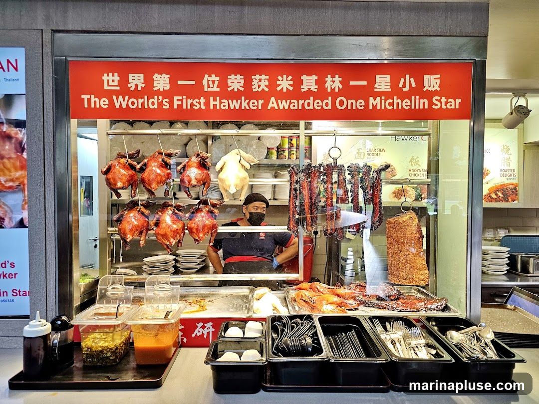 A hawker stall with roasted chickens and pork hanging, and a display of prepared meats. A sign above reads "The World's First Hawker Awarded One Michelin Star". Condiments and cutlery are arranged in front.