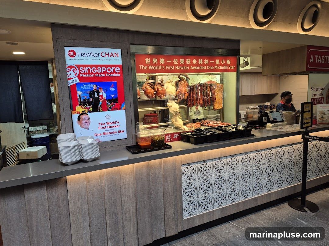 A food stall with roasted chickens and ducks hanging, and a display of prepared food. A screen shows an advertisement for Singapore and mentions Hawker Chan as the first hawker awarded one Michelin star. A staff member is visible behind the counter.
