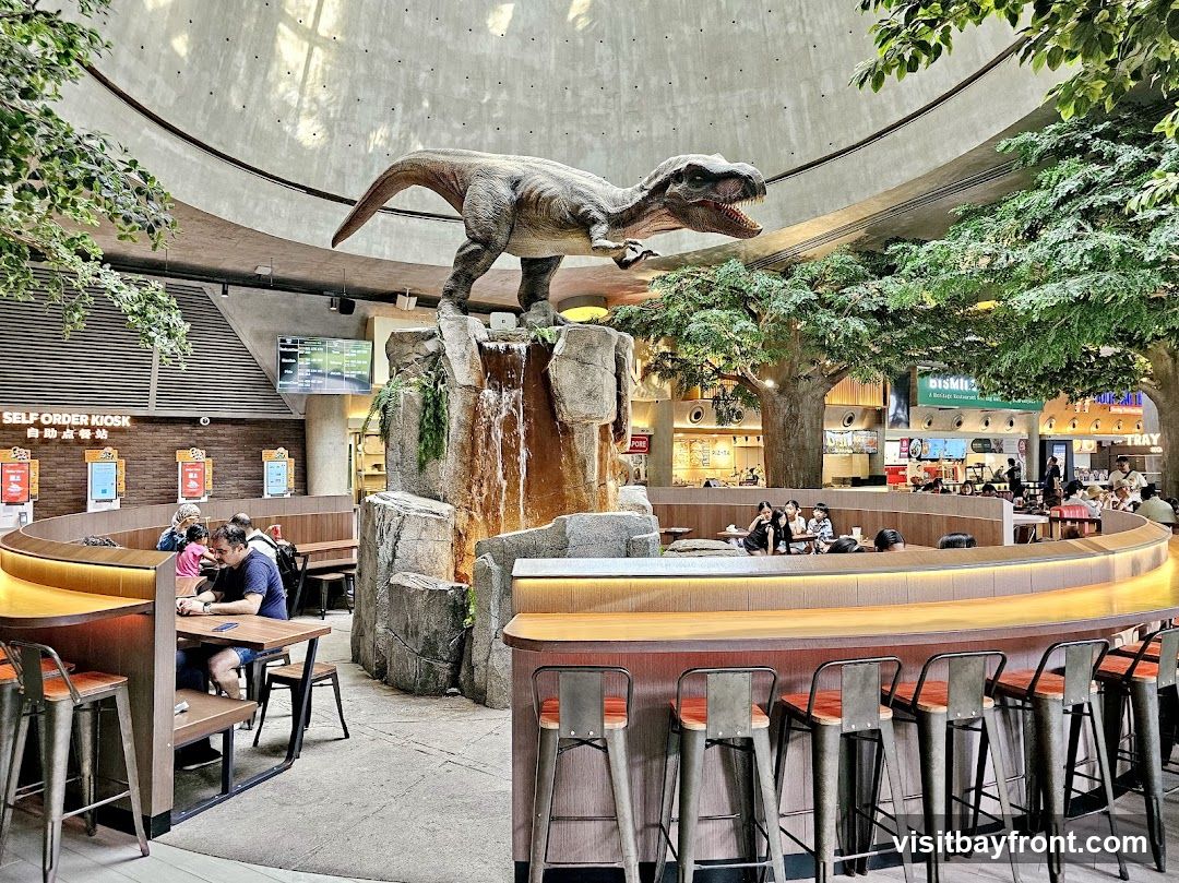 A food court with a large dinosaur statue and a waterfall feature. There are self-order kiosks, seating areas with tables and chairs, and a bar with stools. Several people are dining and ordering food. The space is decorated with artificial trees and has a domed ceiling.