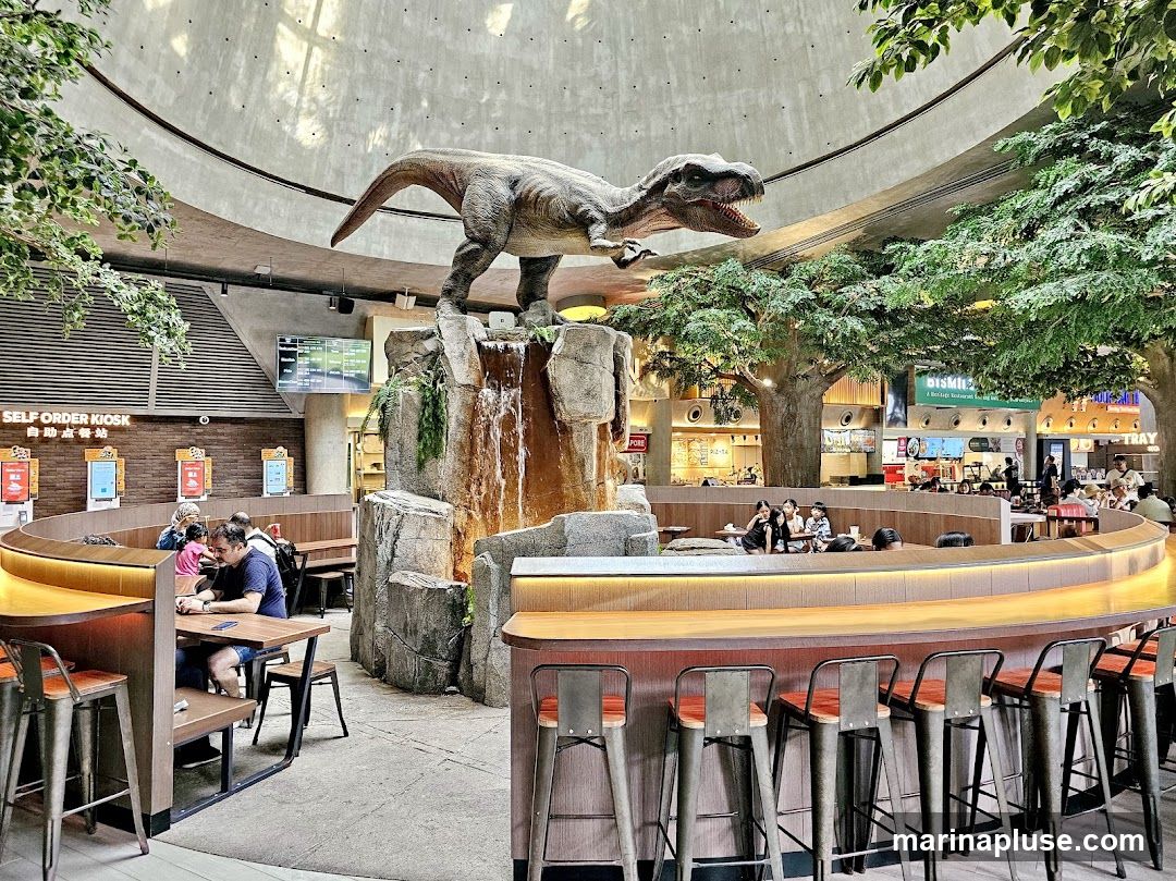 A food court with a large dinosaur statue and a waterfall feature. There are self-order kiosks, seating areas with tables and chairs, and a bar with stools. Several people are dining and ordering food. The space is decorated with artificial trees and has a domed ceiling.