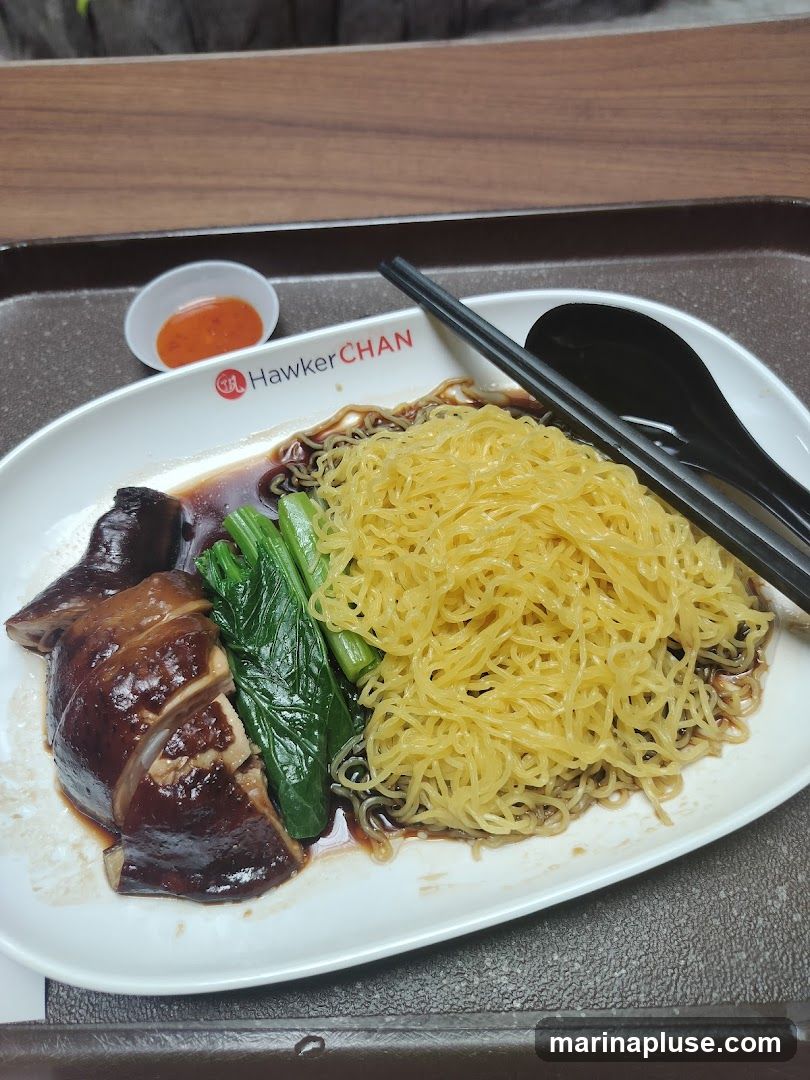 A plate of noodles with chopped braised chicken and green vegetables, served with a small dish of chili sauce. The plate has the Hawker Chan logo.