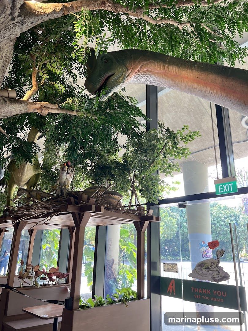 A display featuring realistic dinosaur models, lush artificial foliage, and a seating area. One large dinosaur model extends from the top left of the frame, appearing to eat leaves from a tree. Below, a smaller dinosaur nest display holds several baby dinosaur figures. The scene is set indoors with large windows offering a view of an outdoor area. An 'EXIT' sign is visible above the glass doors, and a 'Thank You, See You Again' sticker with a dinosaur graphic is affixed to the glass.