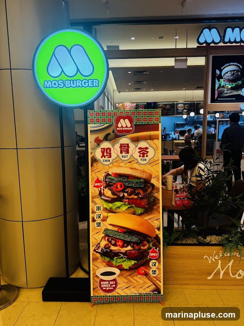 A tall, vertical advertisement for Mos Burger featuring two burgers and Chinese characters. The top burger is labeled as spicy and appears to have chicken, mushrooms, and chili peppers. The bottom burger is also labeled as spicy and has similar ingredients. Below the burgers, there is a small bowl of dark soy sauce with the text "DARK SOY SAUCE will be given." The advertisement is placed in front of a Mos Burger storefront, with a prominent green circular sign displaying the Mos Burger logo and name.
