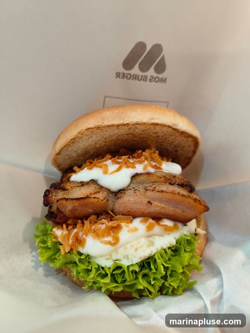 A close-up shot of a burger with a toasted bun, grilled chicken, lettuce, and a creamy sauce, sprinkled with crispy fried onions. The logo of MOS Burger is visible in the background.
