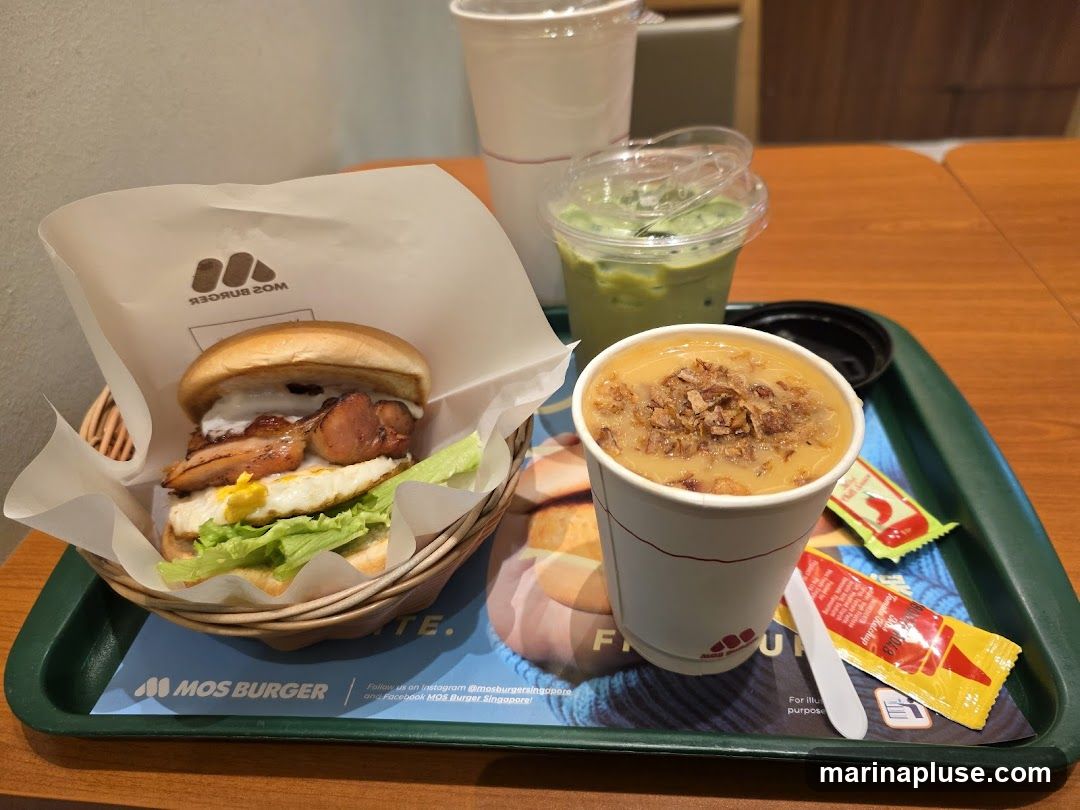 A burger, a cup of soup, and a green drink are on a tray. The burger is in a basket lined with white paper that has the MOS Burger logo. The soup is in a white paper cup and topped with fried onions. The green drink is in a clear plastic cup with a lid. There are also two small packets of condiments and a white plastic spoon on the tray. The tray has a blue mat with the MOS Burger logo and social media information.
