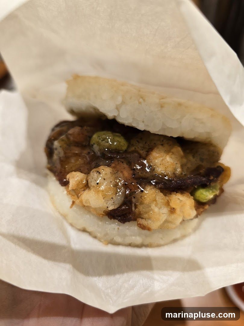 A close-up shot of a rice burger, where the buns are made of pressed rice patties. The filling appears to be a savory mixture with some green elements, possibly edamame, and a glossy sauce. The burger is wrapped in white paper.
