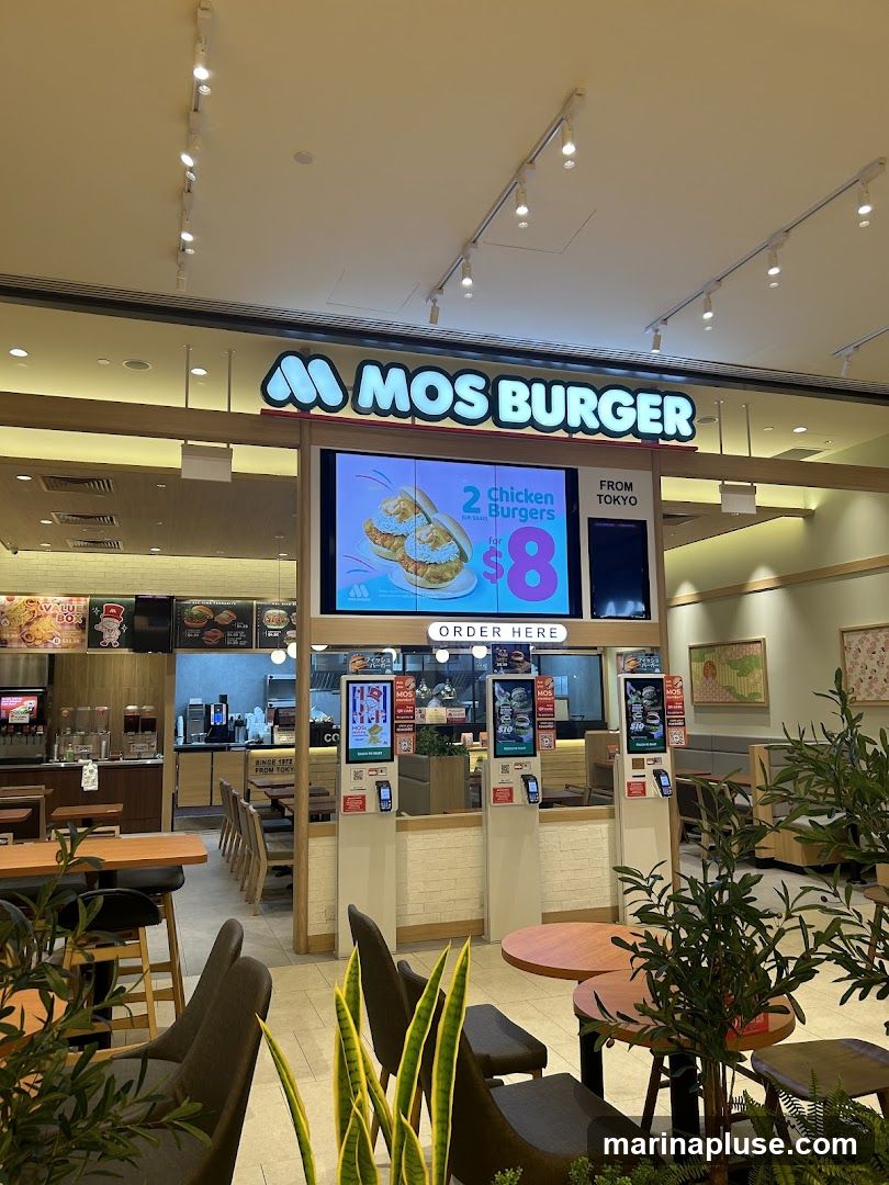 A Mos Burger restaurant interior with a large digital menu board displaying a promotion for 2 chicken burgers for $8. The restaurant features seating areas with tables and chairs, and a counter with ordering kiosks. The brand's logo is prominently displayed above the menu.