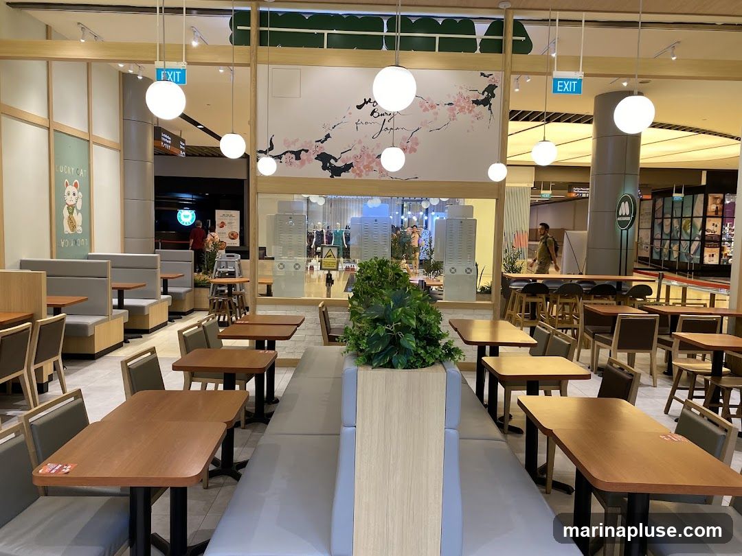 The interior of a restaurant with wooden tables and grey booth seating. A decorative mural of cherry blossoms is on the wall. There are also exit signs and lighting fixtures visible.