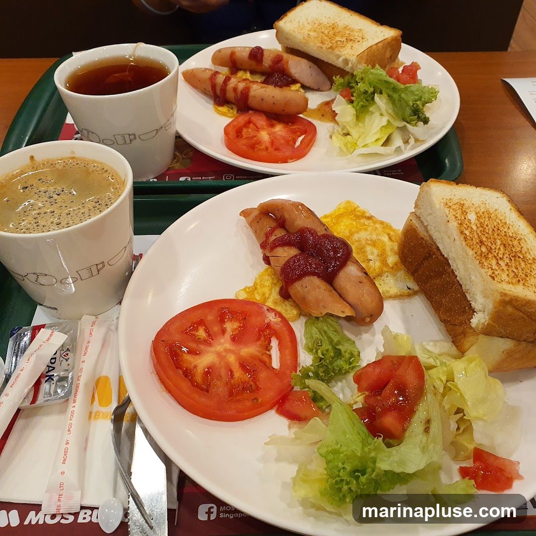 Two plates of breakfast are shown. Each plate contains sausages with ketchup, scrambled eggs, a slice of tomato, a side salad with lettuce and tomato, and toast. Two mugs, one with tea and one with coffee, are also visible. The branding "MOS Burger" is visible on the tray liner and on packaging.