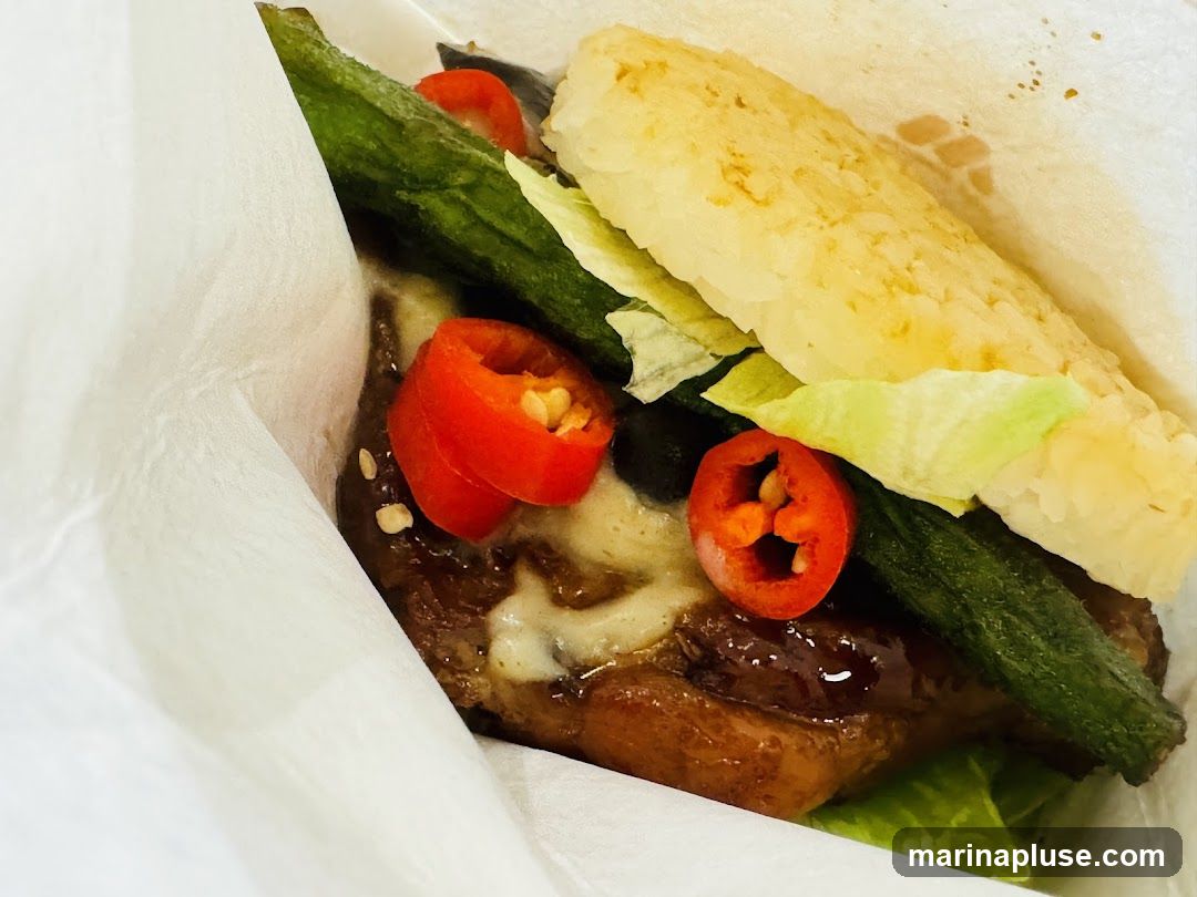 A close-up shot of a rice burger, wrapped in white paper. The burger features a patty topped with melted cheese, sliced red chilies, green beans, and lettuce. The buns appear to be made of pressed rice.