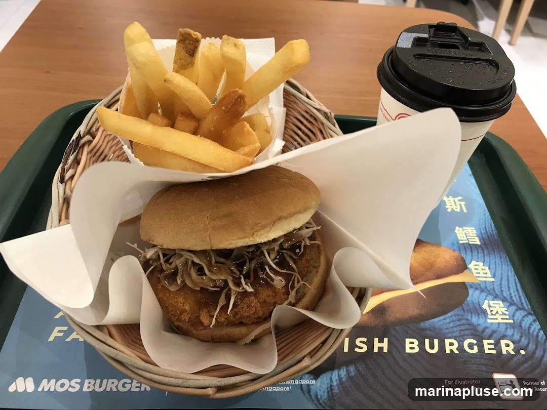 A close-up shot of a MOS Burger meal on a tray. The meal includes a burger, french fries, and a drink in a to-go cup. The burger appears to be a fish burger with a crispy patty, topped with shredded cabbage and sauce. The french fries are golden brown and served in a paper wrapper. The tray liner has the MOS Burger logo and text that reads "FISH BURGER."