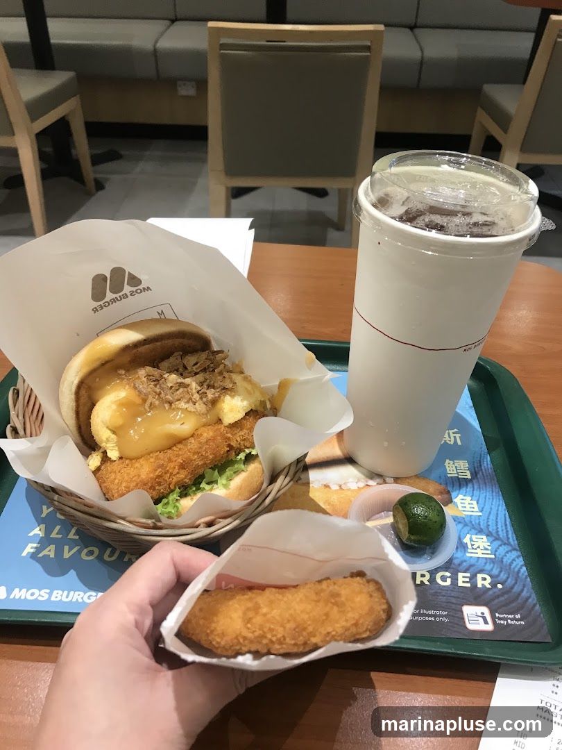 A Mos Burger meal is laid out on a table, featuring a burger in a paper wrapper, a drink in a white cup, and a fried item held in a hand. The burger appears to have a fried patty, lettuce, and a sauce or topping. The drink is a dark liquid with ice. A small lime wedge is also visible. The meal is placed on a green tray with a printed advertisement or menu featuring Chinese characters and the Mos Burger logo.