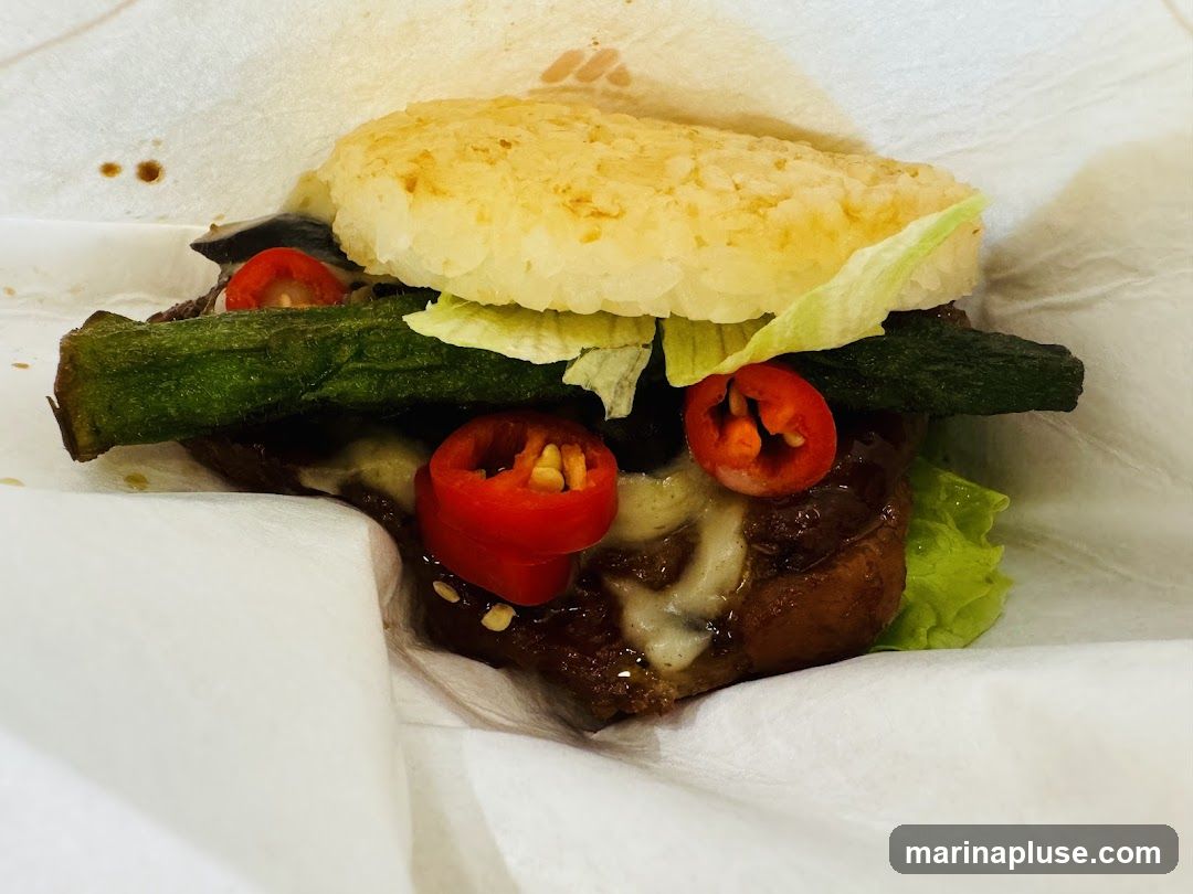 A close-up shot of a burger with rice buns. The burger is filled with lettuce, grilled okra, sliced red chili peppers, and a meat patty with melted cheese. The burger is wrapped in white paper.