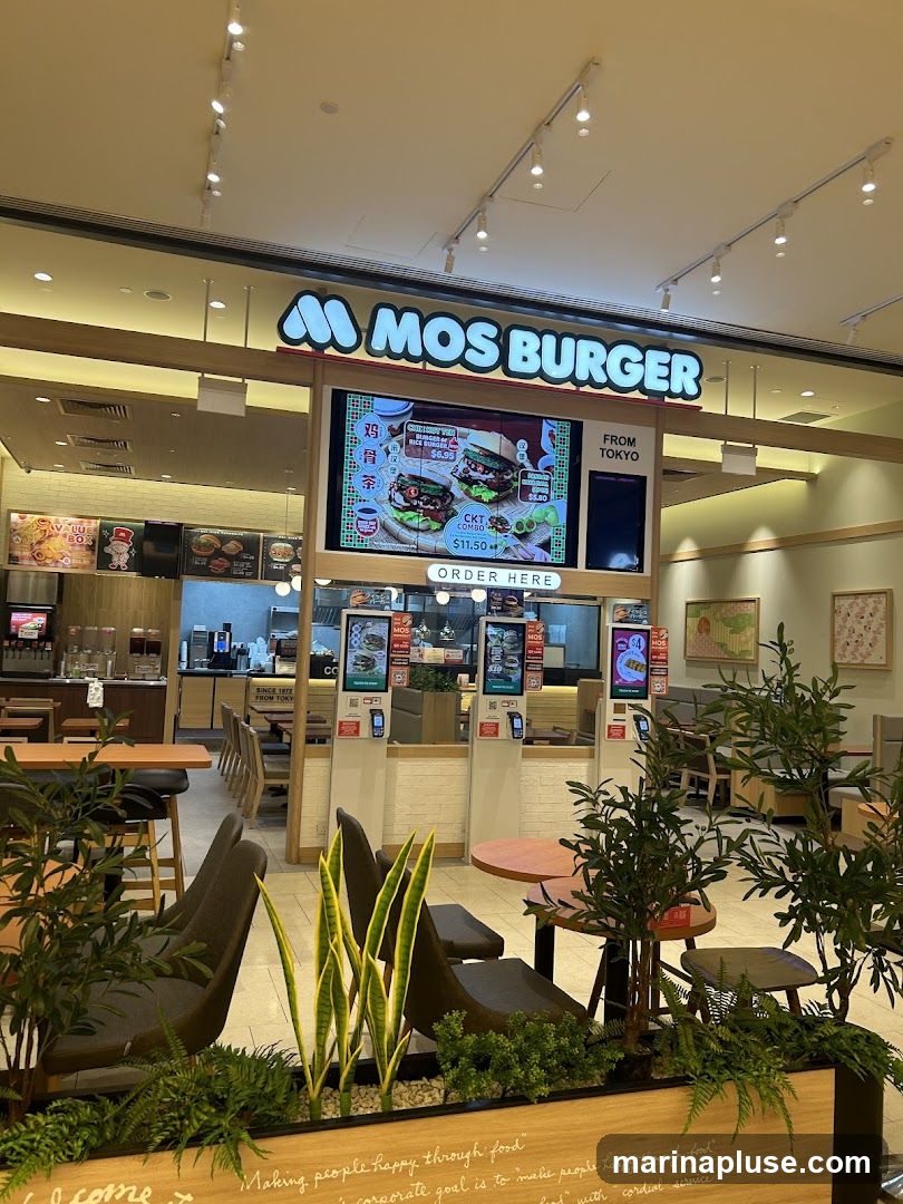 A MOS BURGER restaurant interior with a large digital menu board displaying burgers and combos. The sign above reads "MOS BURGER FROM TOKYO". There are tables and chairs visible, as well as plants in the foreground.