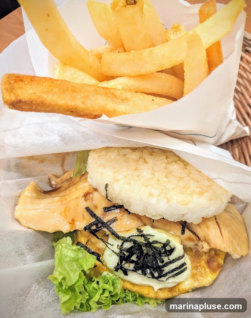 A close-up shot of an onigirazu (rice burger) with a side of french fries. The onigirazu appears to have grilled salmon, lettuce, egg, and nori seaweed, all wrapped in rice and possibly nori. The fries are golden brown and appear to be thick-cut.