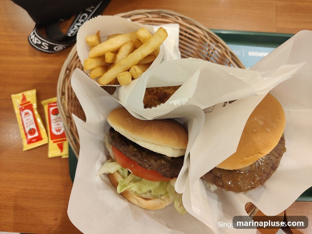 Two burgers and a side of french fries are presented in paper wrappers and a woven basket. Two small packets of ketchup are visible to the left. The food is on a wooden table.