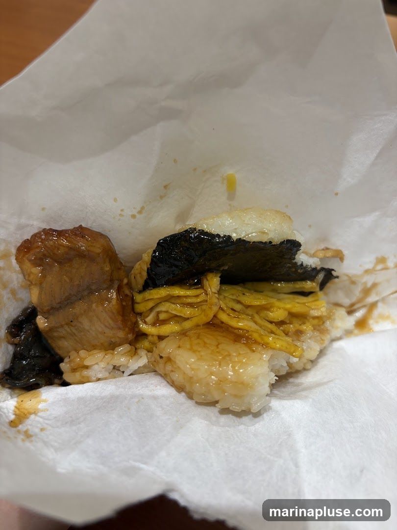 A close-up shot of an onigiri, a Japanese rice ball, wrapped in white paper. The onigiri is filled with grilled eel glazed in a sweet sauce, shredded egg omelet, and a piece of nori seaweed. The rice is sticky and appears to be seasoned.