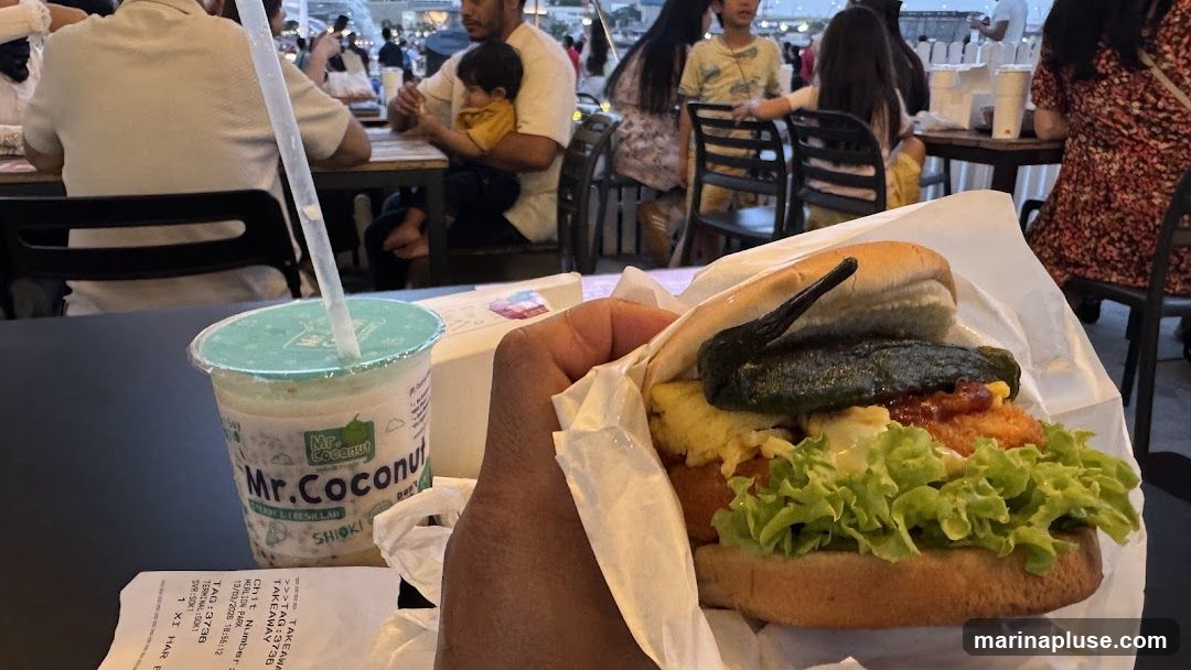 A hand holds a burger wrapped in white paper. Next to it is a cup with a straw, labeled "Mr. Coconut". In the background, people are seated at tables, suggesting an outdoor dining area. A receipt is visible on the table.