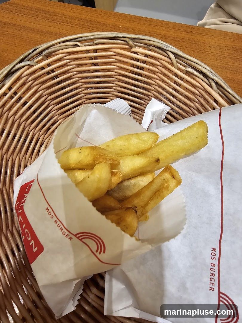 A close-up shot of french fries served in a paper bag with the MOS BURGER logo. The fries are golden brown and appear to be seasoned. The paper bag is white with red accents and text. The fries are placed in a woven basket on a wooden table.