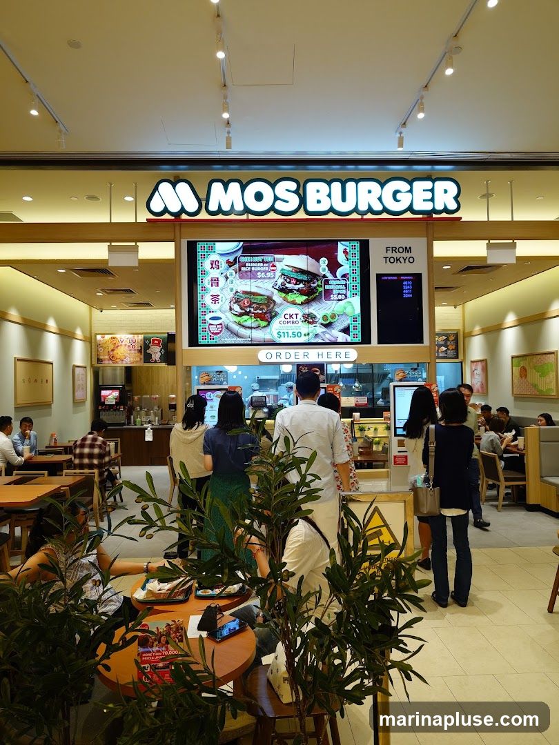 An interior view of a Mos Burger restaurant with a large digital menu board displaying burgers and combos. The "MOS BURGER" logo is prominently displayed above the screen. Customers are seen ordering and dining at tables.