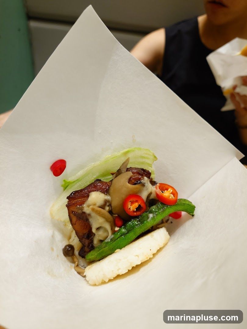 A close-up shot of a dish featuring a piece of pork belly, rice, mushrooms, green beans, and sliced red chili peppers, all wrapped in white paper. A person is visible in the background holding a food item.