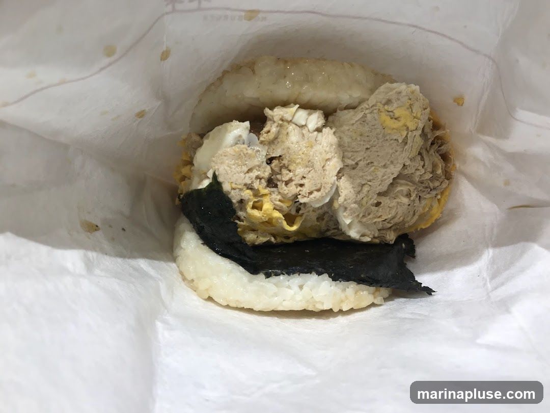 A close-up shot of an onigirazu, a type of Japanese rice sandwich, wrapped in white paper. The sandwich has rice patties on the top and bottom, with a filling of scrambled eggs, shredded meat, and a sheet of nori seaweed in between.
