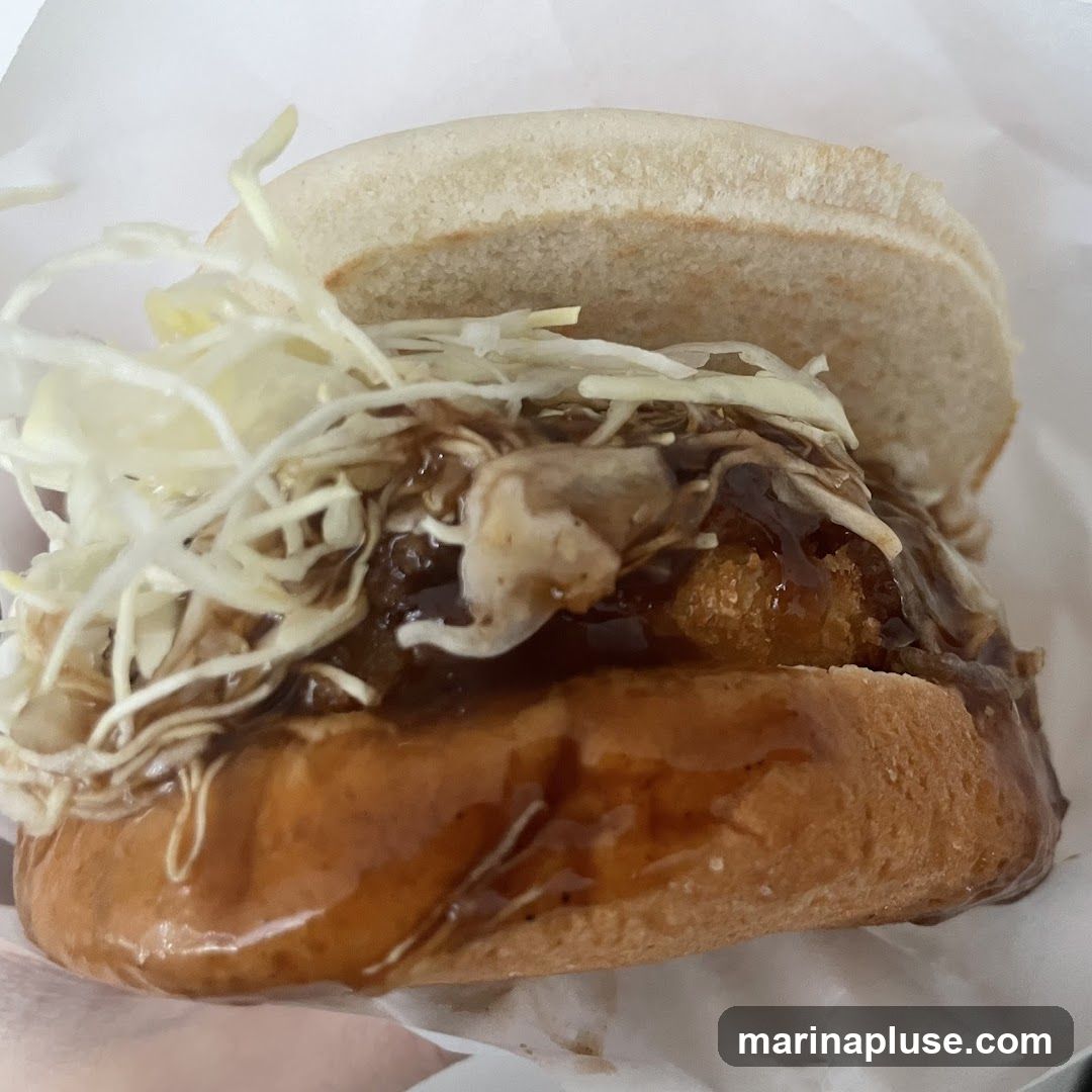 A close-up shot of a burger with a white bun, topped with shredded cabbage and a dark, glossy sauce. The filling appears to be fried and saucy. The burger is wrapped in white paper.