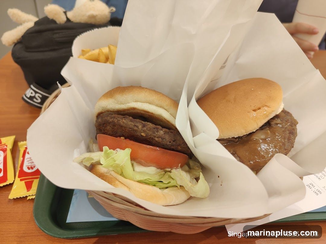 A close-up shot of two burgers and a side of fries served in a basket. The burgers appear to be made with beef patties, lettuce, and tomato. One burger is topped with a brown sauce. Two small packets of ketchup are visible on the left. A stuffed toy is partially visible in the background. The image is dated 2024.12.23.
