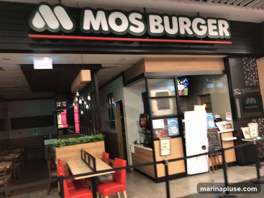The exterior of a MOS BURGER restaurant. The sign above the entrance reads "MOS BURGER" in large white letters with a green outline. The entrance area has a counter with menu boards and seating areas visible inside.