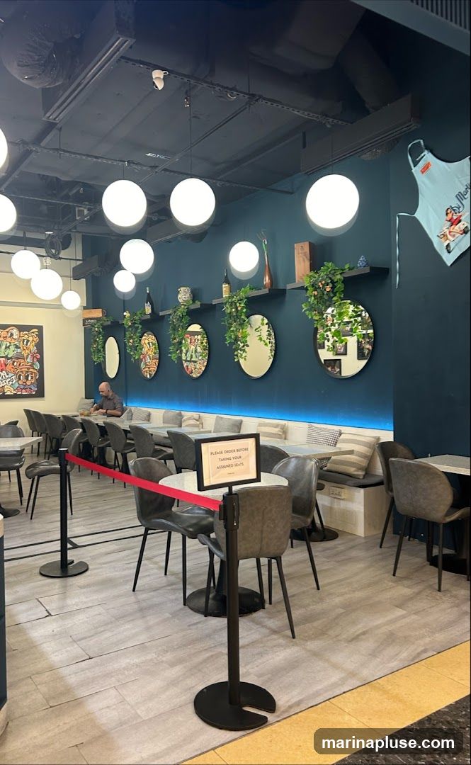 The image shows the interior of a restaurant with a long banquette seating area along a teal-colored wall. Several round mirrors with greenery are mounted on the wall. A man is seated at a table in the background. There are also individual tables and chairs in the foreground. A sign on a stanchion reads "PLEASE ORDER BEFORE TAKING YOUR ASSIGNED SEATS." Large spherical light fixtures hang from the dark ceiling. A colorful framed artwork is visible on the left wall. A light blue apron with a cartoon image is hanging on the right wall.