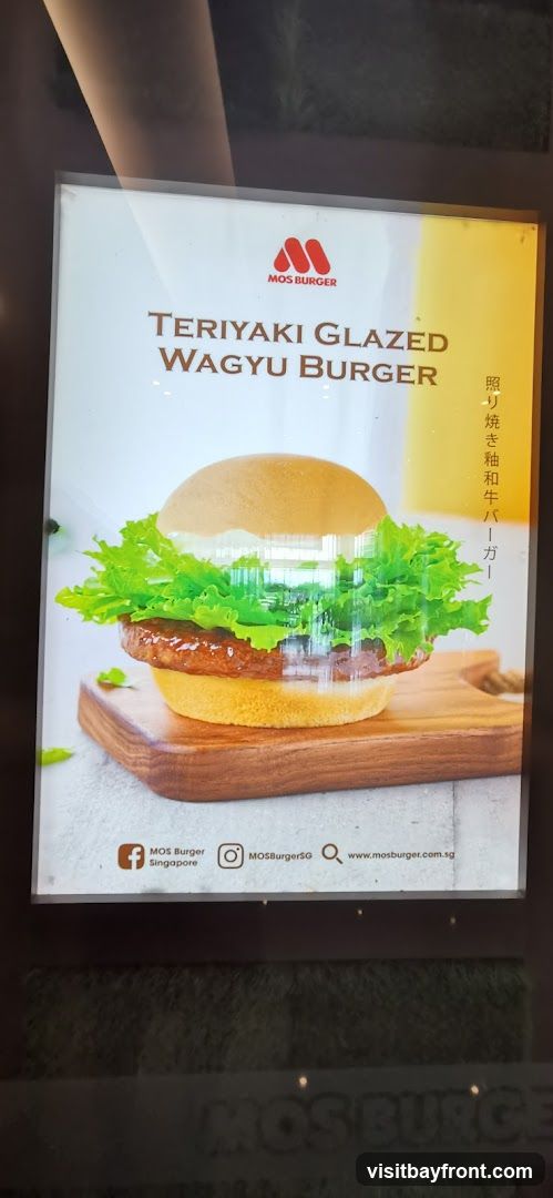 A close-up of a digital advertisement for MOS Burger's Teriyaki Glazed Wagyu Burger. The ad features a large, appetizing image of the burger with lettuce and a thick patty, presented on a wooden board. The MOS Burger logo is visible at the top, along with the product name in English and Japanese. Social media handles and a website address are displayed at the bottom.
