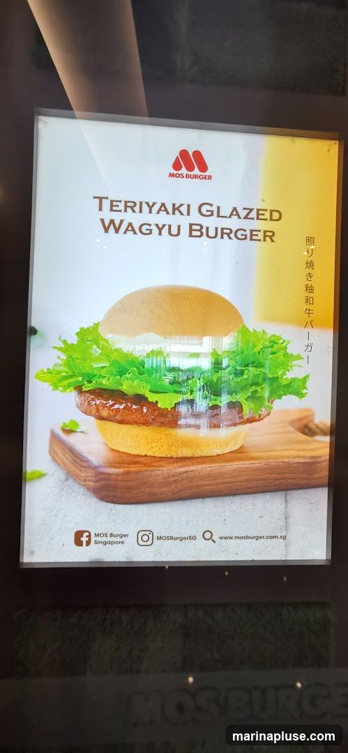 A close-up of a digital advertisement for MOS Burger's Teriyaki Glazed Wagyu Burger. The ad features a large, appetizing image of the burger with lettuce and a thick patty, presented on a wooden board. The MOS Burger logo is visible at the top, along with the product name in English and Japanese. Social media handles and a website address are displayed at the bottom.