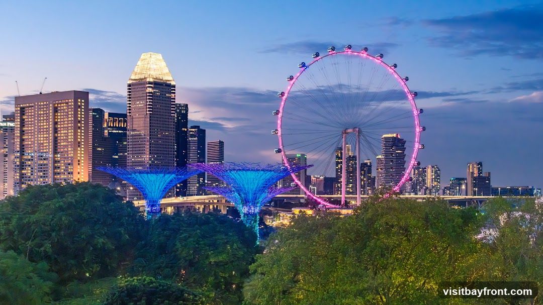 A panoramic view of the Singapore skyline at dusk, featuring the illuminated Gardens by the Bay Supertrees, the Singapore Flyer Ferris wheel, and various skyscrapers. The sky is a gradient of blue and purple, with clouds adding texture. Lush green trees in the foreground frame the cityscape.