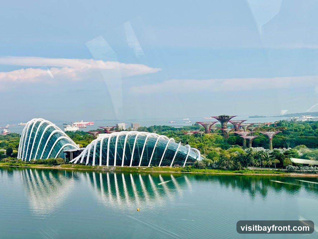 A panoramic view of Gardens by the Bay in Singapore, featuring the iconic Supertree Grove and the Cloud Forest and Flower Dome conservatories. The structures are set against a backdrop of lush greenery, a calm body of water, and a hazy sky with ships in the distance. The architecture of the conservatories is characterized by their curved, glass-paneled roofs supported by white lattice-like structures. The Supertrees, tall vertical gardens, are visible in the mid-ground, resembling futuristic trees. The water in the foreground reflects the sky and the structures, creating a serene and picturesque scene.