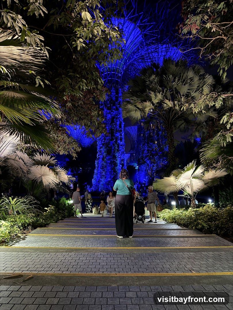 A pathway with stone steps leads up towards a large, illuminated structure in a garden at night. The structure and surrounding trees are bathed in vibrant blue light, creating a surreal and magical atmosphere. Several people are walking up the steps, enjoying the spectacle. Lush foliage frames the scene, with palm trees and other tropical plants visible.