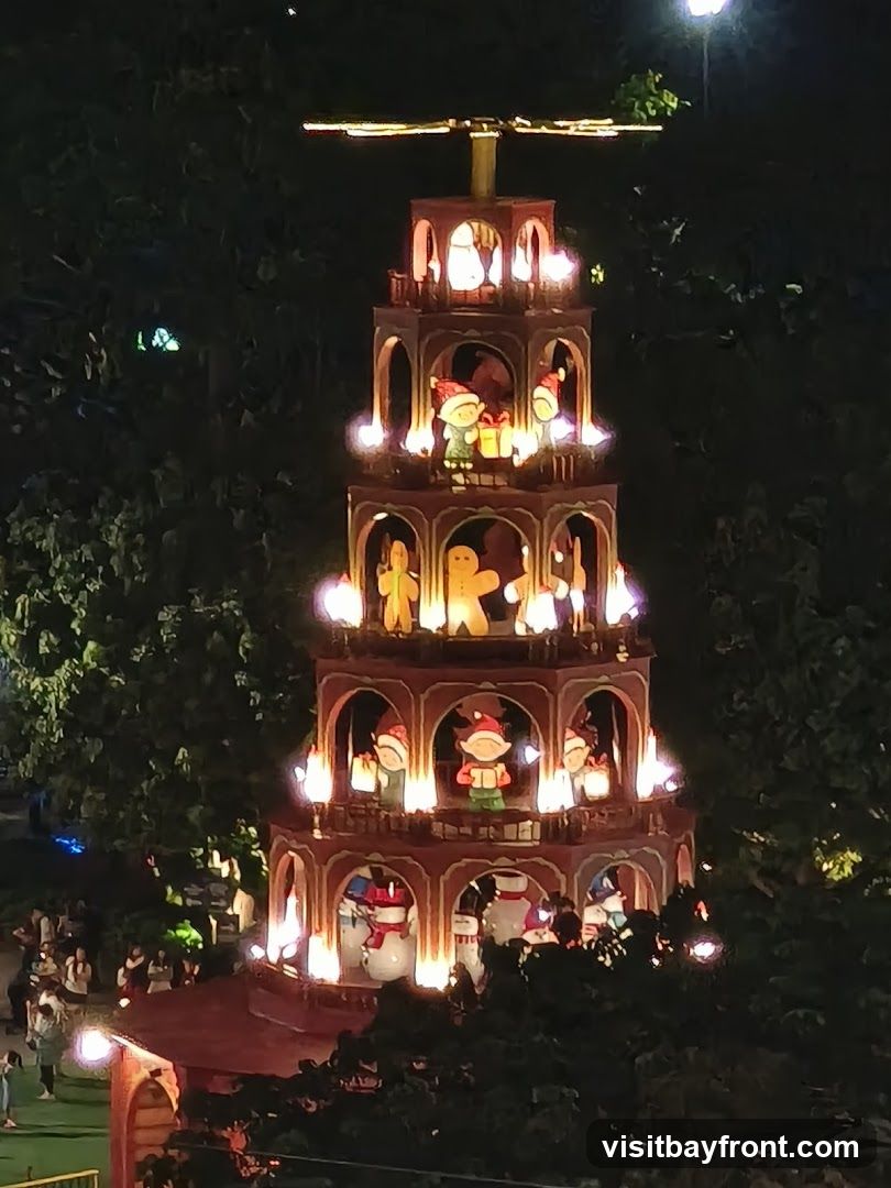 A tall, multi-tiered Christmas pyramid decoration is illuminated at night, surrounded by trees. The pyramid features various festive figures, including elves, snowmen, and gingerbread men, on each level. Lights are placed around the edges of each tier, creating a warm glow. People are visible in the background, suggesting an outdoor event or gathering.