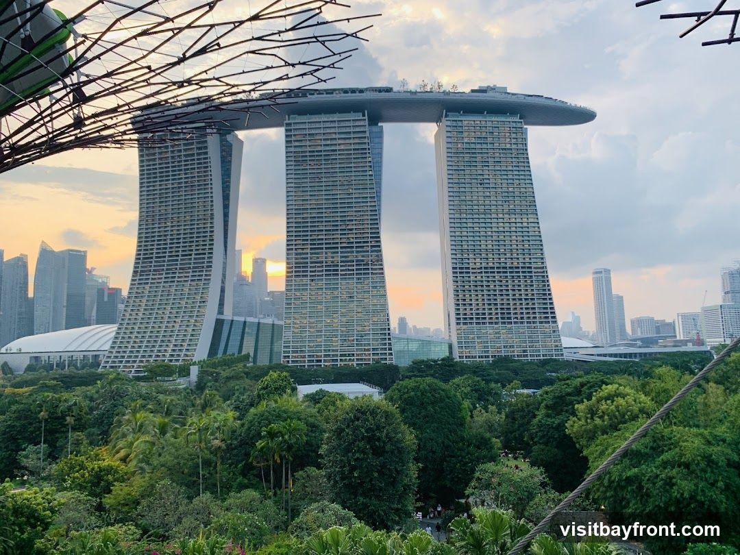 A panoramic view of the Marina Bay Sands hotel and its iconic SkyPark, set against a backdrop of the Singapore skyline at sunset. In the foreground, lush greenery from Gardens by the Bay creates a vibrant contrast with the modern architecture. The sky is filled with soft clouds, illuminated by the warm hues of the setting sun.