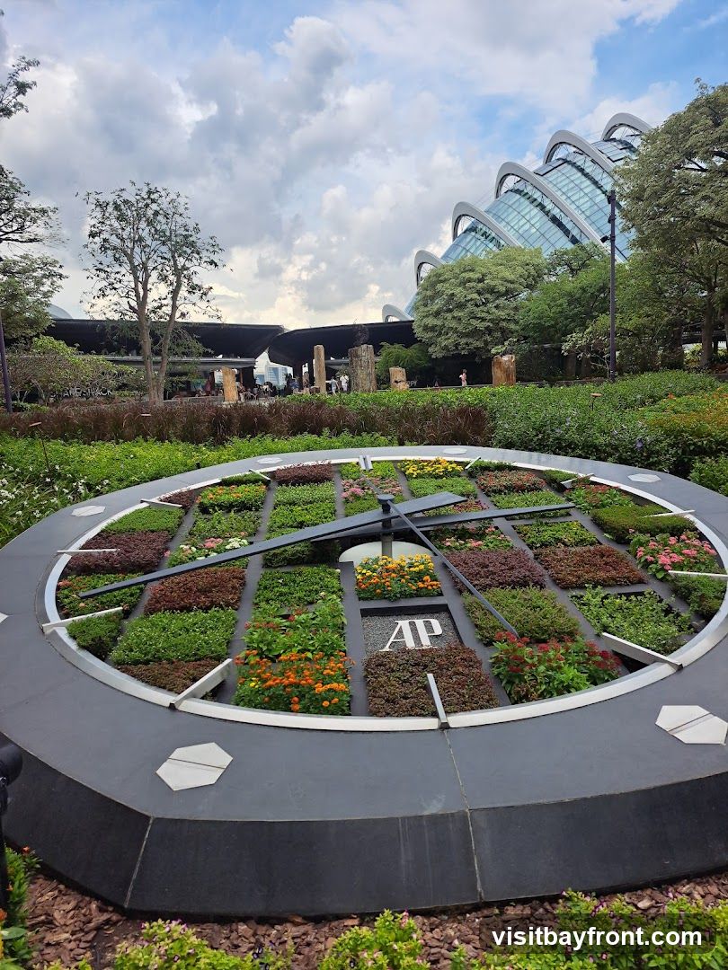 A large, circular clock face filled with various colorful plants and flowers. The clock hands are visible, and the logo 'AP' is displayed in the center. In the background, there are trees and the distinctive architecture of a large dome-shaped building. The sky is cloudy.