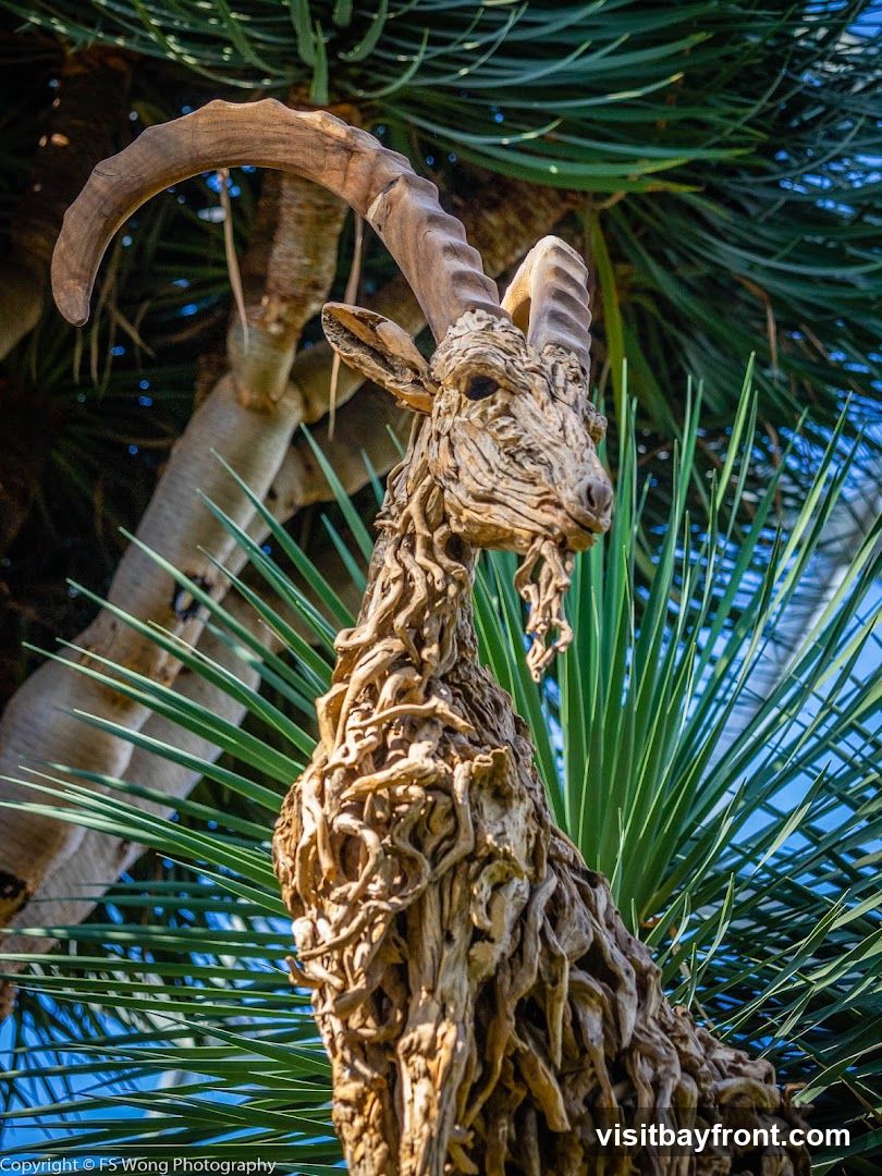A detailed sculpture of an ibex, crafted from weathered wood, is set against a backdrop of lush green palm fronds and a clear blue sky. The sculpture captures the animal's distinctive curved horns and textured body, with individual pieces of wood artfully arranged to form its shape.