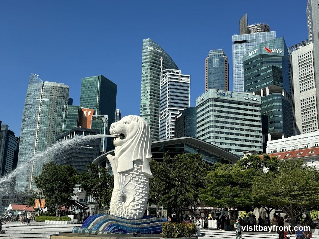 A view of the Merlion statue, a famous landmark in Singapore, with its mouth spouting water. In the background, the modern skyline of Singapore is visible, featuring numerous skyscrapers with various architectural designs and company logos such as CapitaLand, Wework, and MYP. The sky is clear and blue.