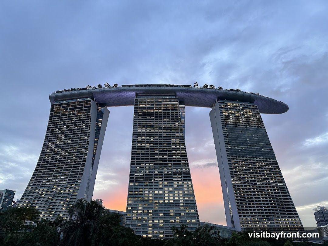 A low-angle shot of the Marina Bay Sands hotel in Singapore during dusk. The sky is filled with clouds, with a gradient of purple and orange hues near the horizon. The three towers of the hotel are illuminated, with many windows lit up. The iconic SkyPark is visible at the top, with trees and lights. The building's unique architecture, with its leaning towers and the cantilevered platform, is prominent. Lush greenery is visible in the foreground.