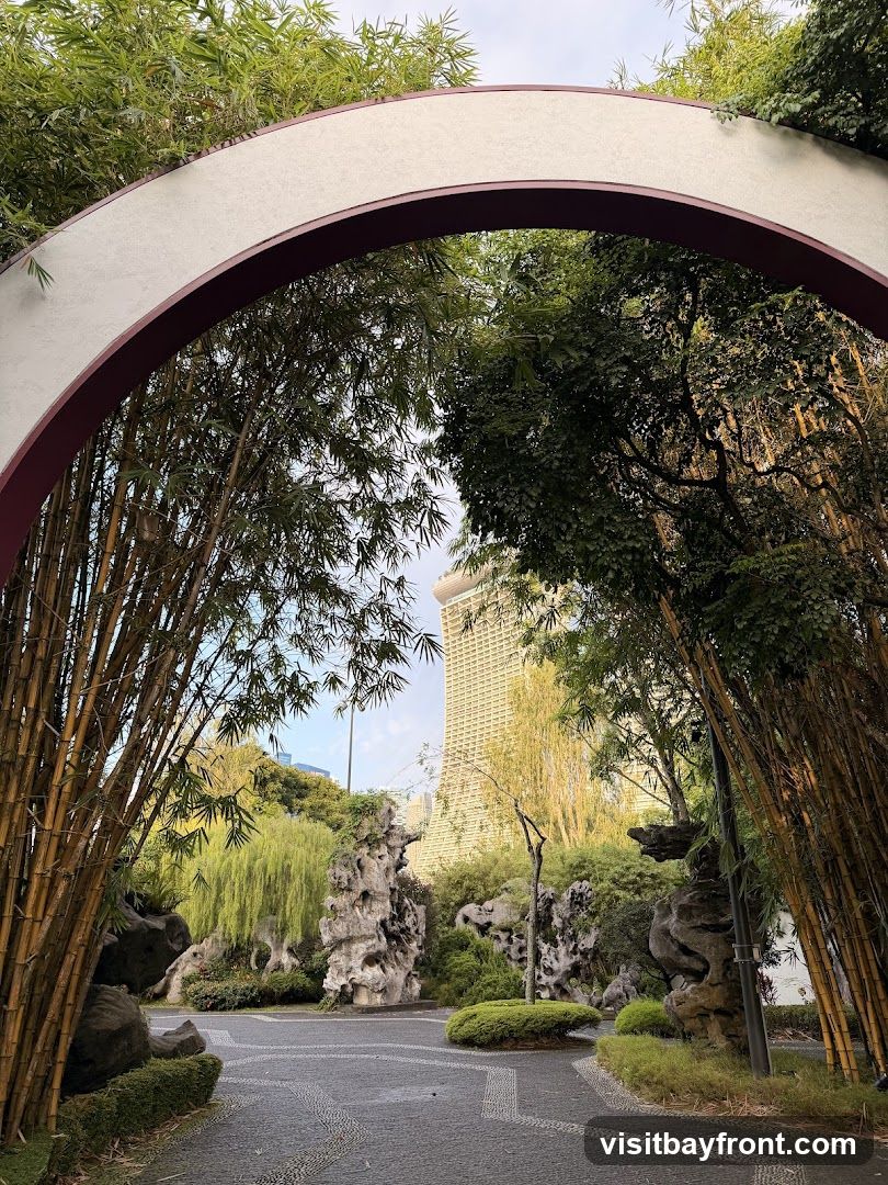 A view through a decorative archway, framed by bamboo stalks and lush greenery. Beyond the arch, a serene garden landscape unfolds with rock formations and weeping willow trees. In the distance, a modern skyscraper stands tall against a pale sky, creating a contrast between nature and urban architecture. The pathway in the foreground is paved with intricate stone patterns.
