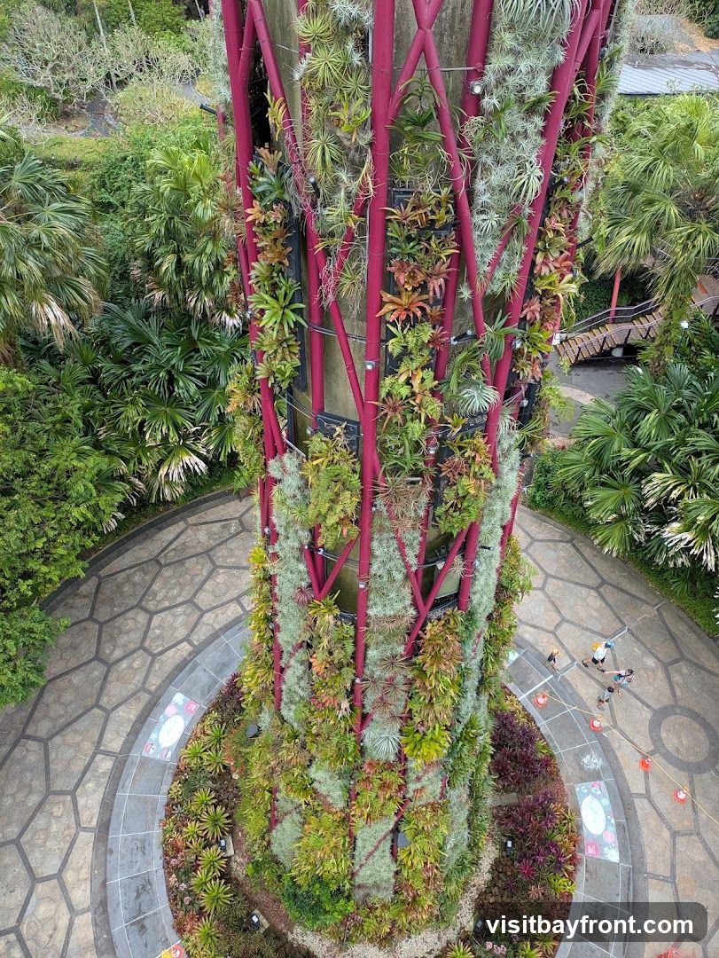 An aerial view of a tall, cylindrical structure covered in a variety of plants, including air plants and bromeliads. The structure has a red, lattice-like framework. Surrounding the base of the structure is a paved pathway with a hexagonal pattern and landscaped areas with more plants. Several people are visible on the pathway below.