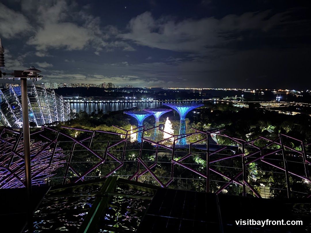 A panoramic night view of Gardens by the Bay in Singapore. The iconic Supertree Grove is illuminated with blue and purple lights, standing tall amidst lush greenery. In the background, the cityscape of Singapore is visible with its buildings and a body of water reflecting the city lights. The foreground features a geometric metal structure with purple accents, possibly part of an observation deck or walkway. The sky is dark with scattered clouds.