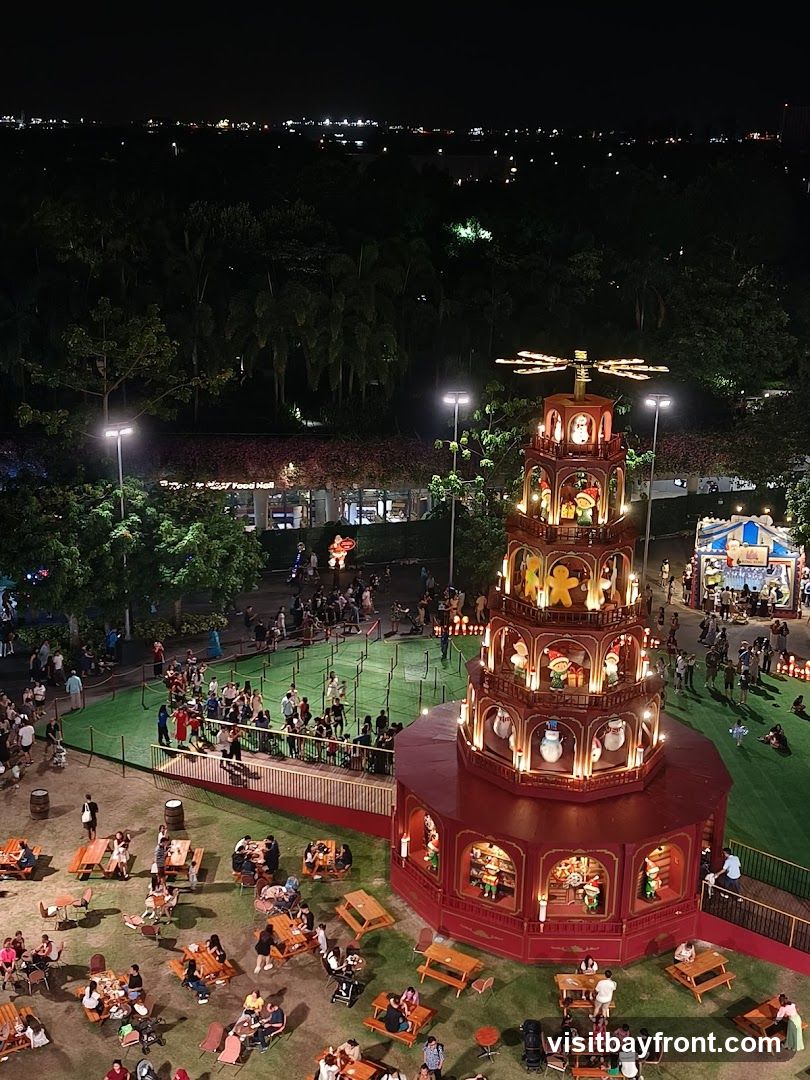 An aerial view of a bustling Christmas market at night. A large, multi-tiered, illuminated Christmas pyramid stands prominently in the center, decorated with festive figures like gingerbread men, snowmen, and Santa Claus. Surrounding the pyramid is a green lawn with numerous picnic tables and chairs occupied by people enjoying the atmosphere. In the background, there are more stalls, lights, and trees, with a cityscape visible in the distance.