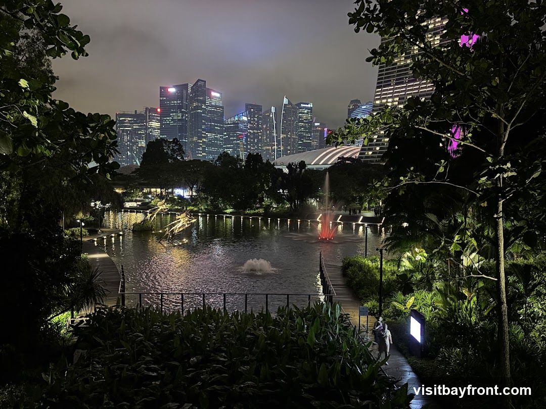 A scenic view of a park at night, with a calm lake reflecting the lights of a modern city skyline in the background. Fountains add a dynamic element to the water, and lush greenery surrounds the pathways. The scene is illuminated by artificial lights, creating a vibrant and atmospheric ambiance.
