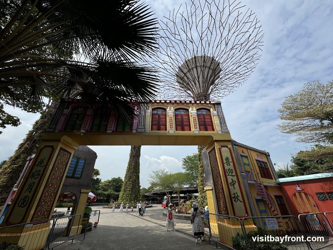 An archway decorated to resemble traditional buildings with colorful windows and doors, set against a backdrop of the Supertree Grove at Gardens by the Bay. Palm fronds frame the top of the image. Several people are walking through the area.