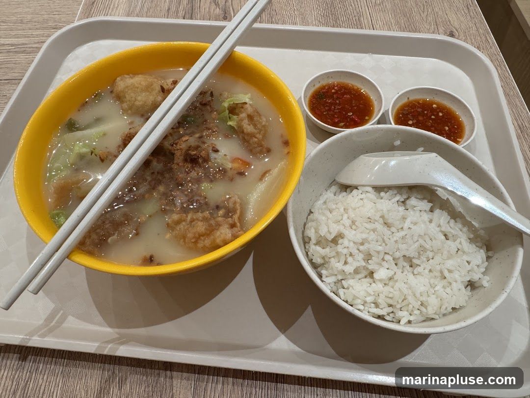 A bowl of fish soup with fried fish pieces, vegetables, and crispy toppings, served with a bowl of white rice, two small bowls of chili sauce, and chopsticks on a tray.