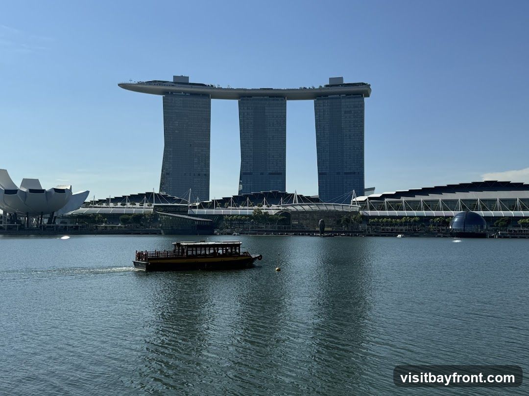 A wide shot of the Marina Bay Sands complex in Singapore, with its iconic three towers connected by a sky-high platform. In the foreground, a traditional wooden boat with a yellow roof sails across the calm blue water. The sky is clear and bright blue.